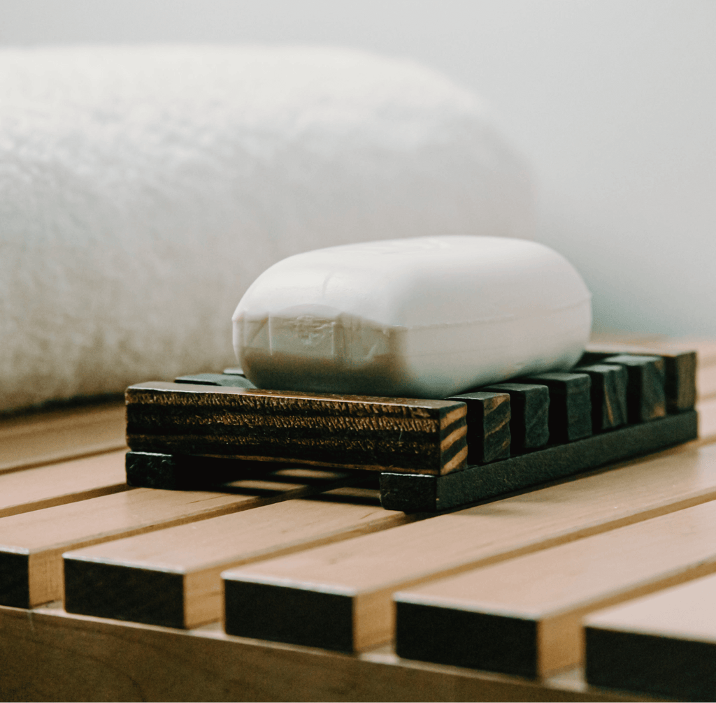 A (Sample) Coconut Bar Soap rests on a wooden soap dish atop a slatted surface, with a rolled white towel in the background.