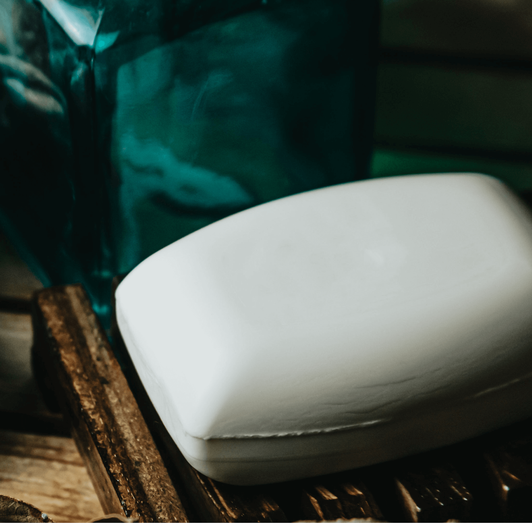 The (Sample) Coconut Bar Soap, a white handmade bar, sits on a wooden soap dish next to a teal glass container with a softly blurred background.