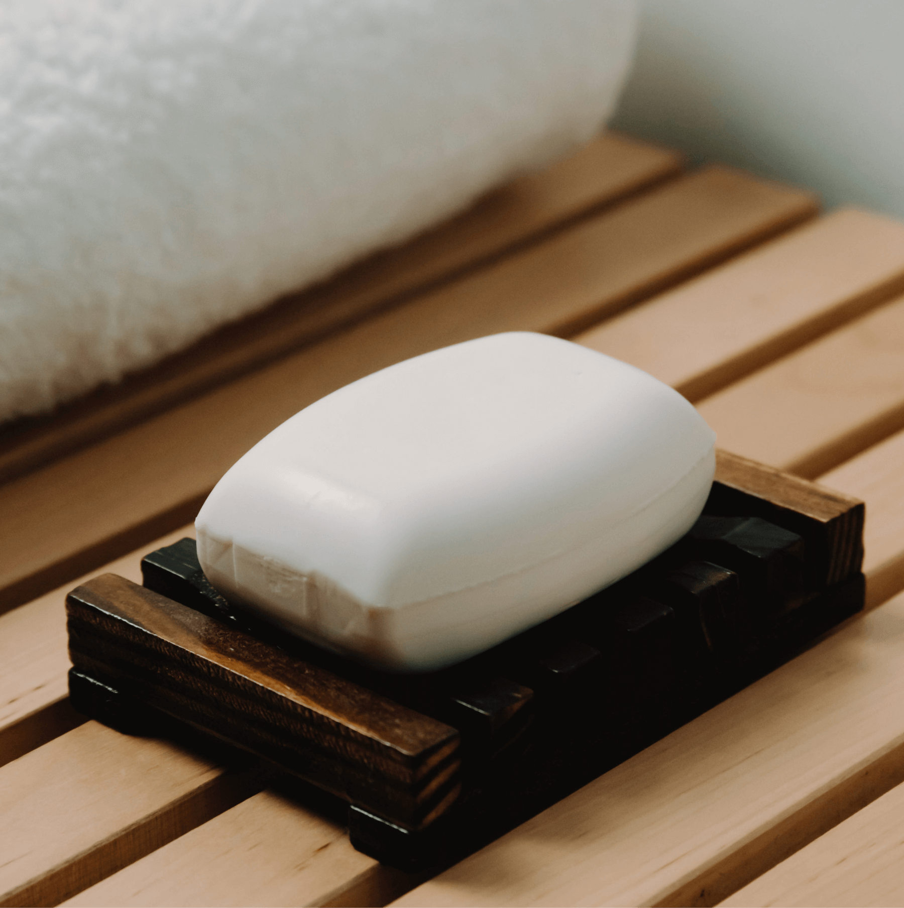 A (Sample) Coconut Bar Soap sits on a dark wood soap dish atop a light wooden slatted surface, with a white towel in the background.